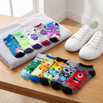 A set of colorful low-cut socks with cartoon monster prints, arranged on a wooden table next to a gray towel and white sneakers.