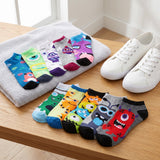 A set of colorful low-cut socks with cartoon monster prints, arranged on a wooden table next to a gray towel and white sneakers.