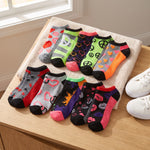 Eight pairs of low-cut ankle socks in assorted colorful patterns are arranged on a beige towel resting on a wooden table, with white sneakers partially visible at the side.