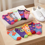A set of colorful novelty ankle socks displayed on a beige towel and tabletop, styled with white sneakers and a cream towel in a bright indoor setting. The socks feature retro designs with hearts, peace signs, flowers, and words like 'LOVE', 'PEACE', and 'JOY'.