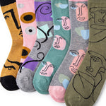 Six pairs of patterned crew socks arranged in a fan shape on a white background. Each sock features a stylized minimalist face illustration in different colors and patterns, including yellow, pink, purple, teal, gray, and olive green.