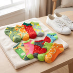 A set of colorful low-cut socks with fruit prints (mango, orange, red strawberry, watermelon, pineapple, and green banana patterns) arranged on a white towel on a wooden table next to a pair of white canvas sneakers.