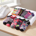 A set of colorful low-cut socks arranged on a white towel on a light wood table, next to a pair of white sneakers. The socks feature various patterns including stripes, hearts, smiley faces, and text.