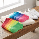A set of ten colorful ankle socks arranged in a fan shape on a cream towel, placed on a wooden bench next to a pair of white canvas sneakers. The socks come in red, orange, yellow, green, light green, purple, blue, and teal.
