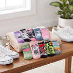 A set of ten colorful cat-themed ankle socks arranged on a beige towel and wooden table, styled with white sneakers, a mug, and a potted plant in a bright indoor setting.