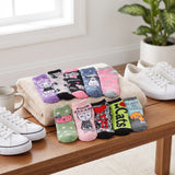 A set of ten colorful cat-themed ankle socks arranged on a beige towel and wooden table, styled with white sneakers, a mug, and a potted plant in a bright indoor setting.