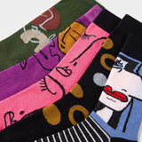 Four pairs of patterned crew socks fanned out on a white background, featuring abstract face designs in green, purple, pink, and black, with yellow, red, and blue accents.
