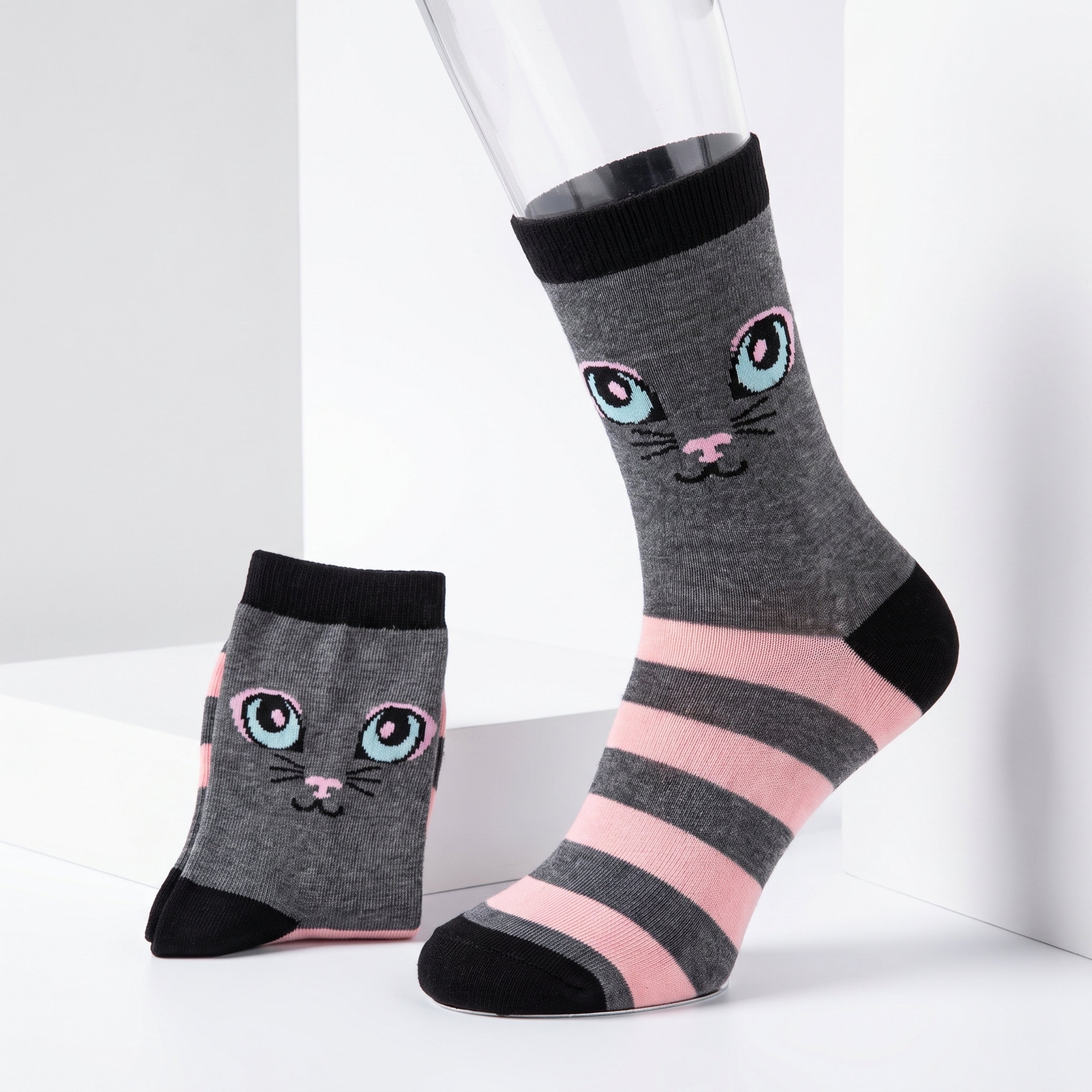 A pair of gray crew socks with a cute cartoon cat face and pink and black stripes, displayed on a white studio background with one sock on a mannequin leg and the other standing beside it.