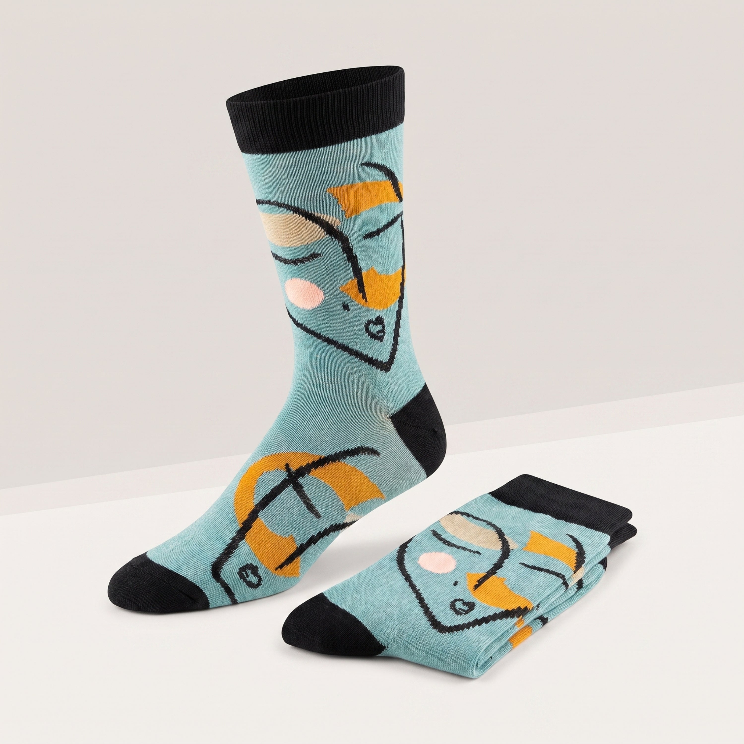 A pair of light blue crew socks with a stylized face graphic in black, yellow, and beige, featuring black heels, toes, and ribbed cuffs, shown on a clean white and light gray background.