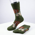 A pair of olive green crew socks with a cartoon mushroom and white line-art pattern, displayed on a white surface with one sock on a clear mannequin leg and the other folded.