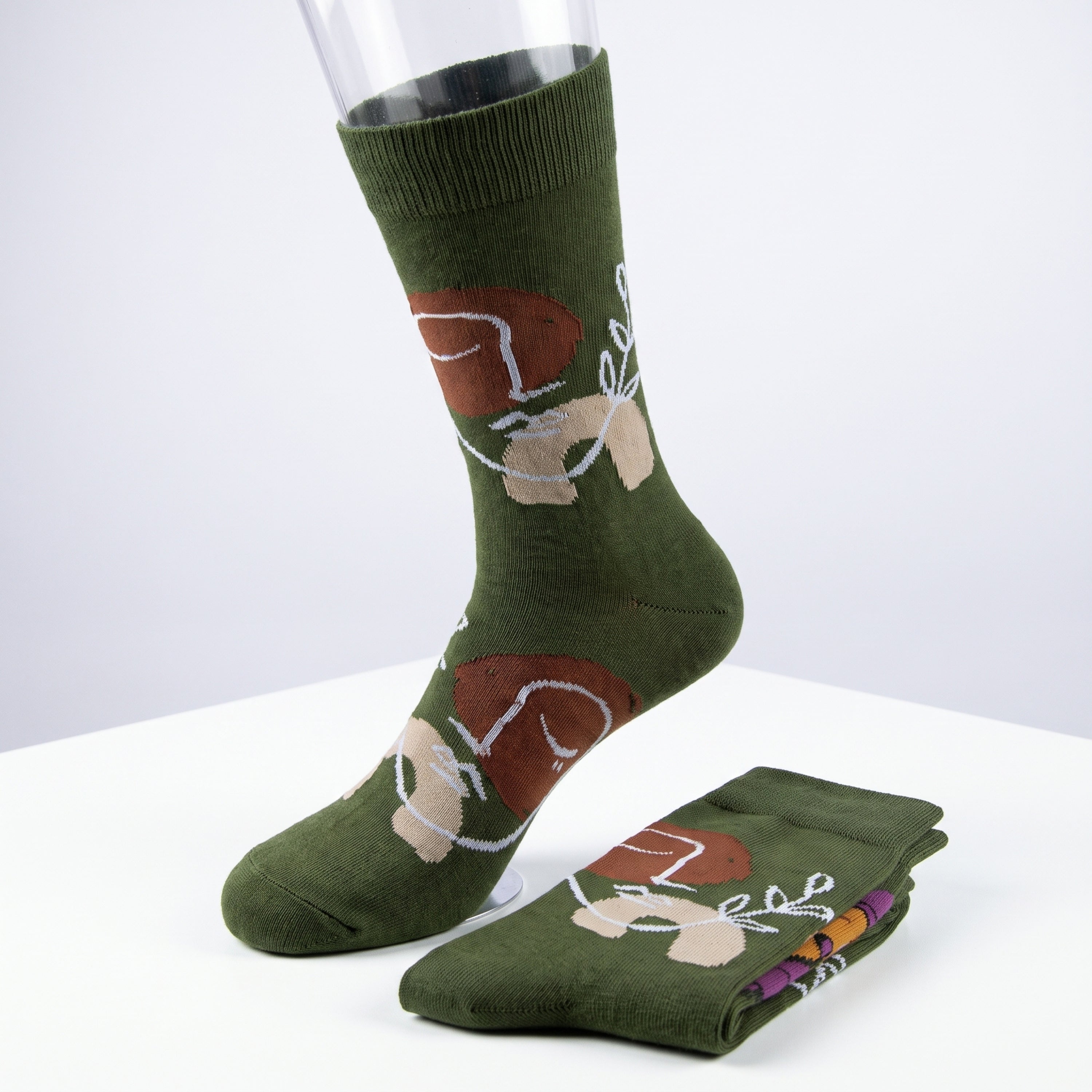 A pair of olive green crew socks with a cartoon mushroom and white line-art pattern, displayed on a white surface with one sock on a clear mannequin leg and the other folded.