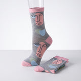 A pair of gray crew socks with a minimalist face illustration pattern in pink, light blue, and beige, shown on a white studio background with one sock on a mannequin leg and the other folded.