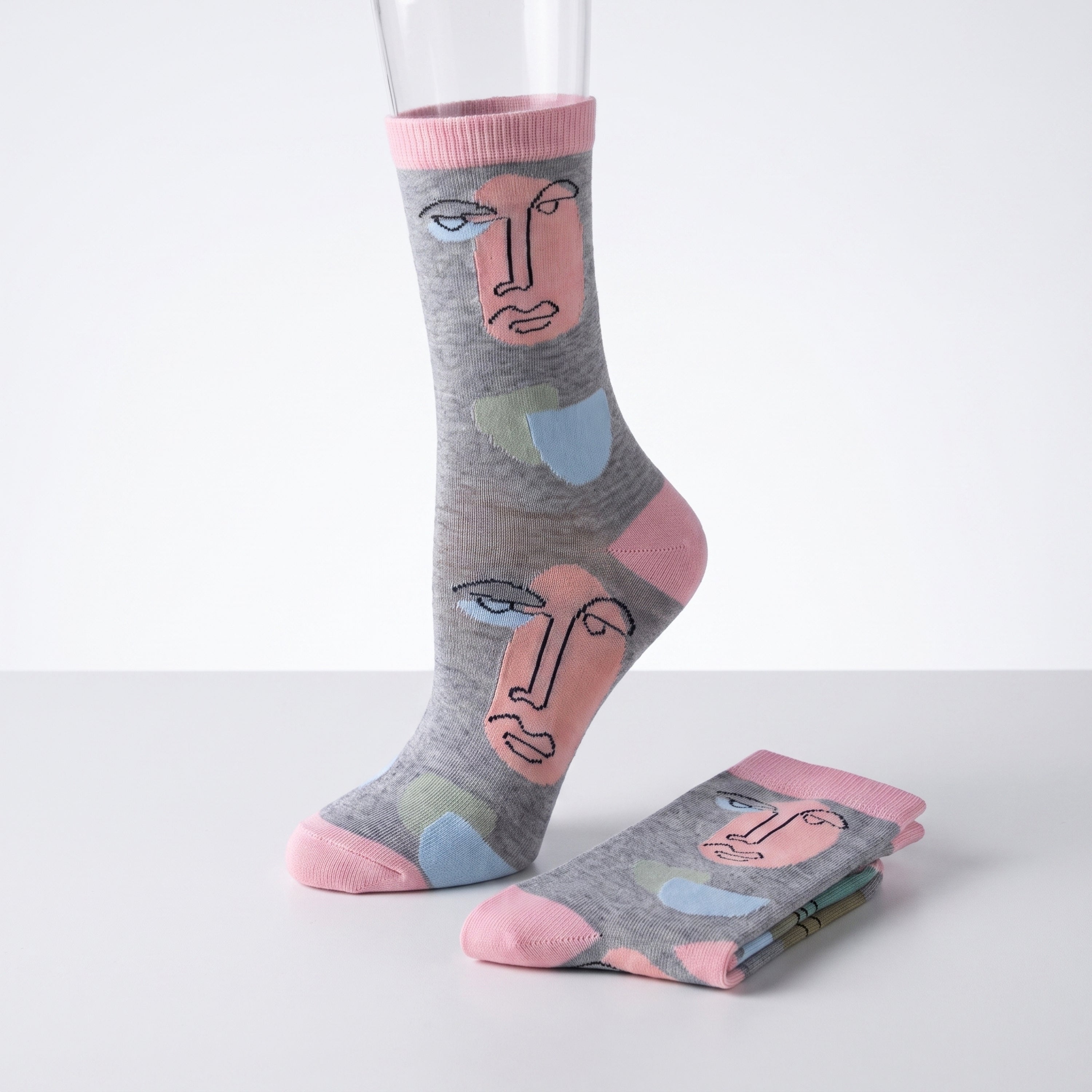 A pair of gray crew socks with a minimalist face illustration pattern in pink, light blue, and beige, shown on a white studio background with one sock on a mannequin leg and the other folded.