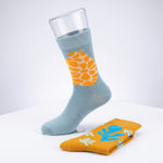 A light blue crew sock with a yellow-orange giraffe print panel is shown on a white surface with a clear glass prop at the top. A folded matching sock in yellow-orange with blue and white leaf-like prints rests in front.