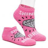 A pair of bright pink ankle socks displayed on a clear mannequin leg against a white background. The socks feature a graphic of a gray diamond with black rays and the words 'SHINE BRILLIANT' in white.