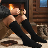 A person sitting by a fireplace wearing black ribbed knee-high socks, holding a mug, with brown lace-up boots on a rug in a rustic cabin setting.