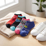 Four pairs of printed ankle socks arranged on a beige towel on a wooden surface, with white sneakers and a potted plant in the background. The socks feature colorful designs including red, black, light blue, pink, gray, and blue pairs with text and graphic prints.