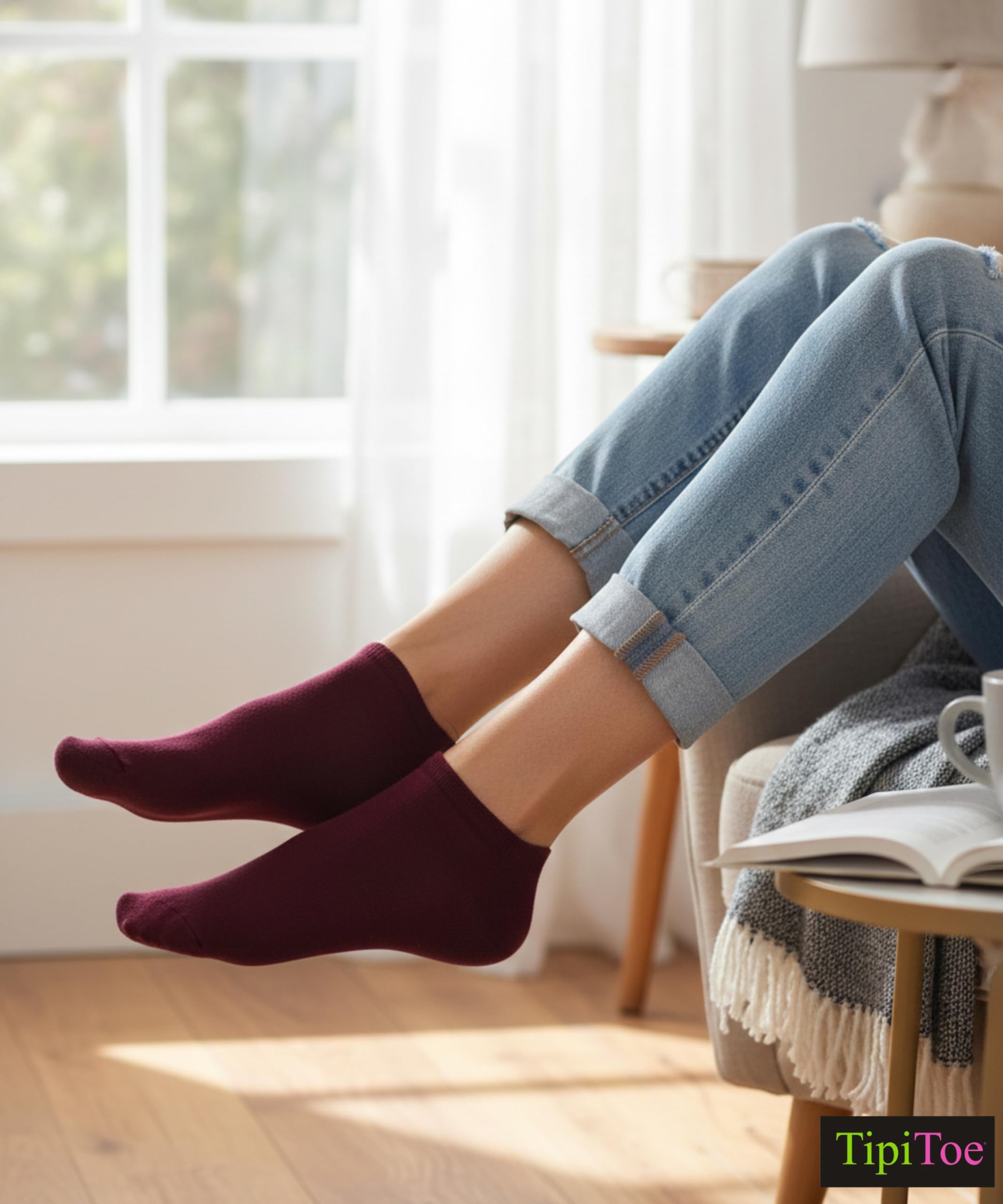 Person wearing maroon socks and light blue jeans sitting on a chair with a book and mug in a cozy room.