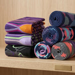 A set of folded and rolled patterned socks displayed on a light wood shelf. The socks feature various colorful geometric and striped patterns in purple, blue, pink, orange, green, and black.