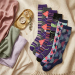 Five pairs of patterned crew socks arranged on a cream blanket with folded pastel textiles and a small gold tray as styling props. The socks feature mixed patterns including argyle, stripes, and abstract floral shapes in purple, pink, blue, green, gray, black, and coral tones.