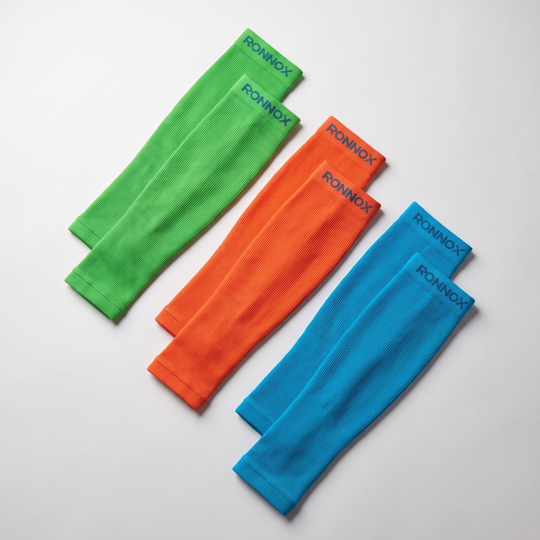 Three pairs of ribbed arm warmers in bright green, orange, and blue, laid out diagonally on a light gray background. Each pair features the brand name 'RONNOX' printed in dark blue on the cuff.