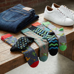 A styled product display showing a variety of colorful patterned crew socks draped over a wooden bench, alongside folded blue jeans and white sneakers on a wooden table.