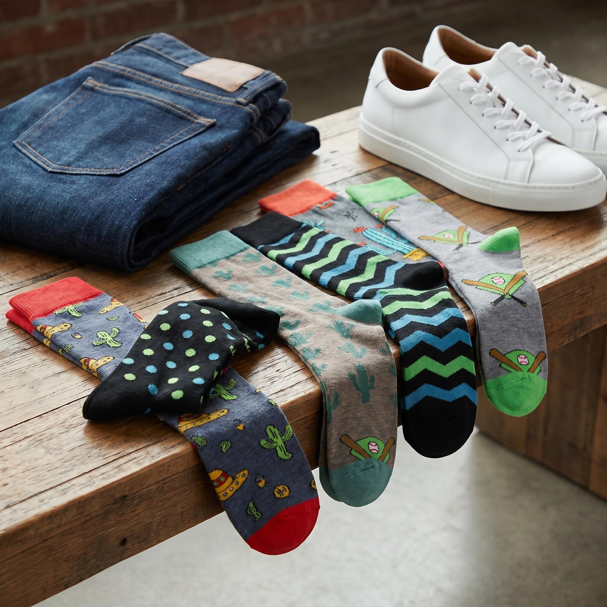 A styled product display showing a variety of colorful patterned crew socks draped over a wooden bench, alongside folded blue jeans and white sneakers on a wooden table.