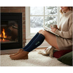 Person sitting by a fireplace wearing navy knee-high socks with a logo, holding a mug and reading a book.