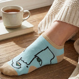 Person wearing a light blue sock with a cat design on a wooden floor next to a cup of coffee and a book.