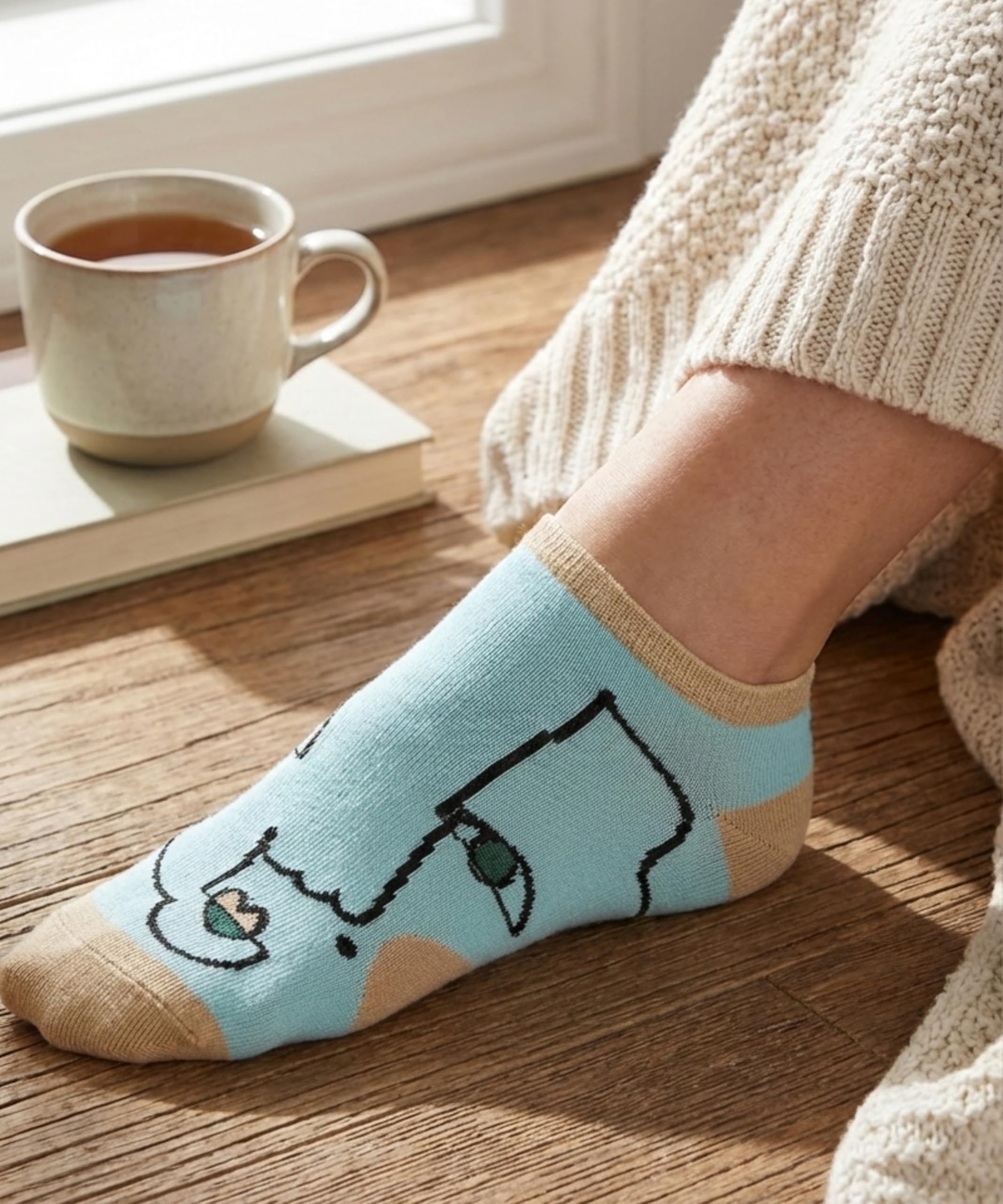 Person wearing a light blue sock with a cat design on a wooden floor next to a cup of coffee and a book.