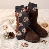 A pair of dark gray knit socks with a beige and brown dog pattern, styled inside brown suede boots on a white cable-knit blanket with wooden snowflakes and a pinecone.
