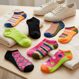 A set of colorful low-cut ankle socks arranged on folded white towels next to a pair of off-white canvas sneakers on a light wood surface. The socks feature various patterns including stripes, polka dots, argyle, and solid colors.