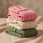 Three folded knit cardigans stacked on a soft beige fabric surface: a dusty pink one on top, a cream one in the middle, and an olive green one at the bottom. Each cardigan has visible wooden buttons.