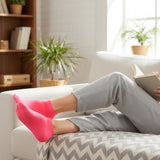 Person relaxing on a couch with pink socks and a book, surrounded by plants and a bookshelf.