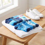 A set of six children's ankle socks with blue ocean and whale patterns is displayed on a white towel on a wooden table, next to a pair of white canvas sneakers.