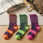Three pairs of colorful striped socks on a wooden surface with a plaid blanket and compass in the background.