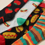 Four pairs of novelty socks arranged diagonally, featuring colorful food-themed patterns including tomatoes, burgers, hot dogs, and striped cheese slices.