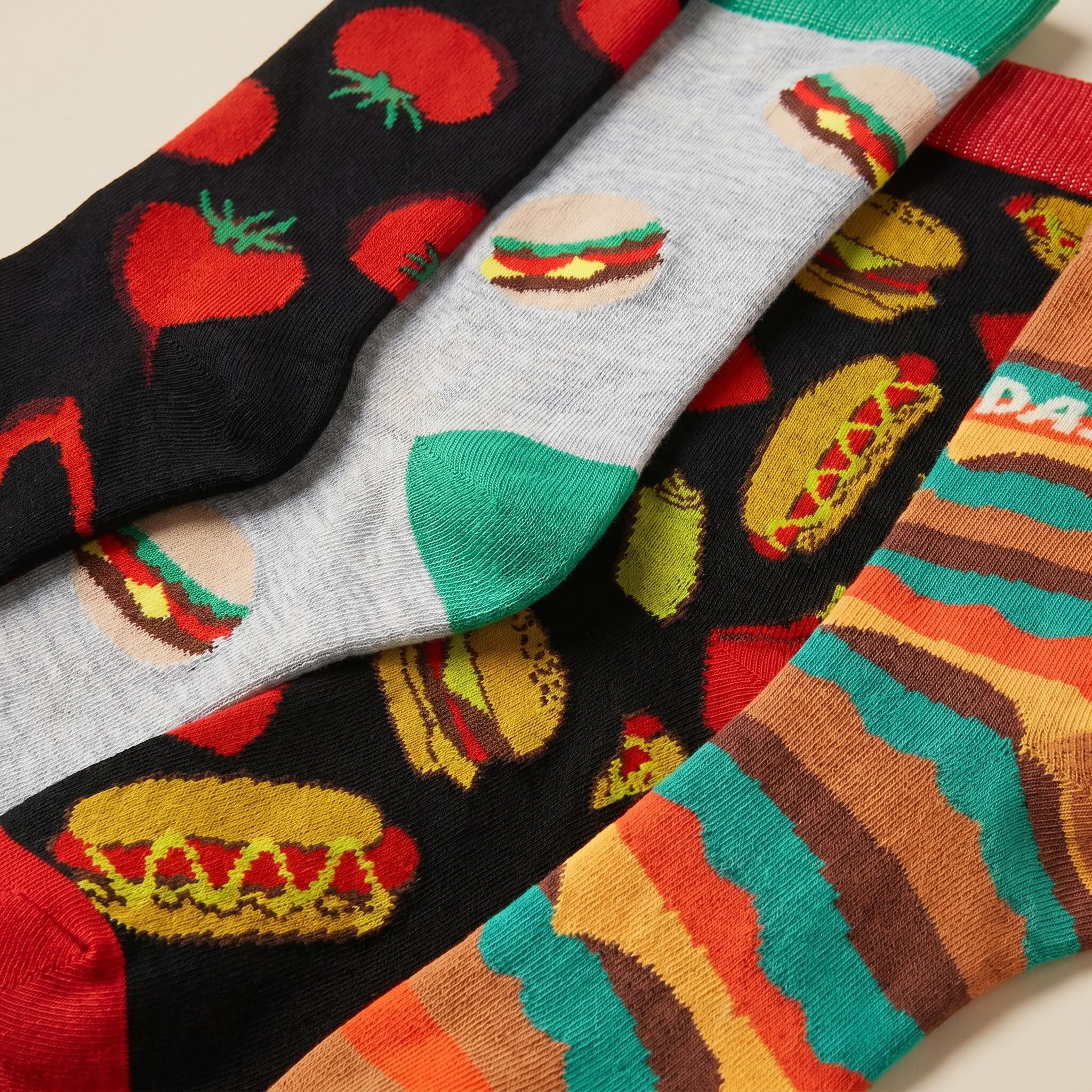 Four pairs of novelty socks arranged diagonally, featuring colorful food-themed patterns including tomatoes, burgers, hot dogs, and striped cheese slices.