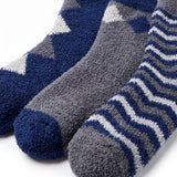 Three plush fleece socks in blue, gray, and white, arranged diagonally on a clean white background. One sock features a geometric star pattern, while the other two display wavy stripes.
