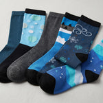 Five pairs of patterned crew socks arranged in a fan shape on a light gray background. The socks feature various winter and abstract designs in shades of blue, teal, gray, and black.