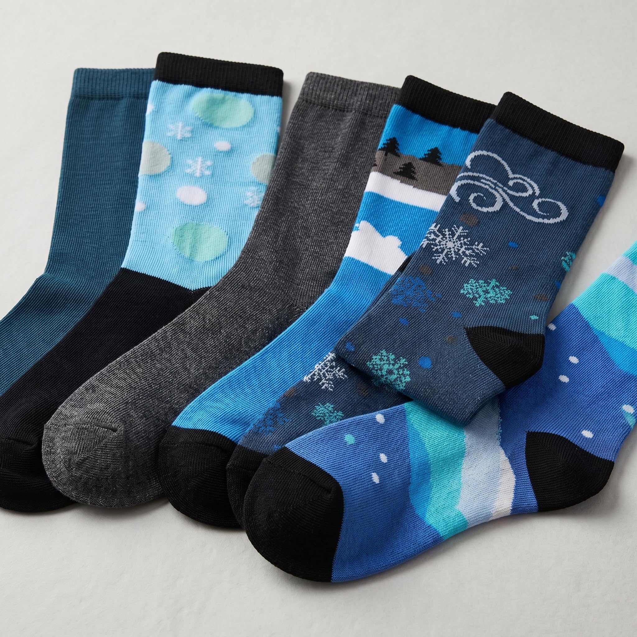 Five pairs of patterned crew socks arranged in a fan shape on a light gray background. The socks feature various winter and abstract designs in shades of blue, teal, gray, and black.