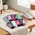 A set of ten colorful graphic crew socks arranged on a white towel on a wooden table, with a pair of white canvas sneakers nearby in a bright, modern room setting.