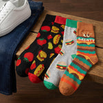 Four novelty crew socks with food-themed patterns are displayed on a wooden bench next to a pair of blue jeans and a white sneaker. The socks feature various designs including tomatoes, burgers, hot dogs, and stripes with the word 'DAINTY'.