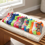 A set of colorful low-cut ankle socks arranged in a row on a folded cream towel resting on a wooden bench, with white sneakers and a window in the background.