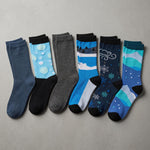 Six pairs of crew socks arranged in a row on a light gray textured background. The socks feature various winter-themed designs including snowflakes, clouds, mountains, and bubbles in shades of blue, gray, black, and teal.
