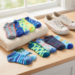A set of colorful patterned ankle socks arranged on a wooden table next to a folded cream towel and a pair of off-white canvas sneakers. The socks feature various designs including stripes, tropical leaves, fish, and starfish in shades of blue, green, yellow, and gray.