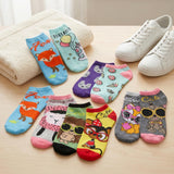 A set of eight colorful low-cut socks with cartoon animal and food illustrations, arranged on a light wood surface next to a folded cream towel and white sneakers.
