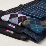 A stack of folded knit socks in multiple colors and patterns, including argyle, stripes, and small embroidered motifs, arranged on a light fabric background.