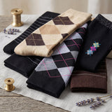 A stack of folded knit socks in multiple patterns and colors, including argyle and striped designs, arranged on a light fabric surface with decorative spools and lavender sprigs.