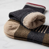 A stack of folded knit socks on a light marble surface. The top sock is dark charcoal gray with a soft beige fleece-like cuff and a red and cream striped ribbed top edge.